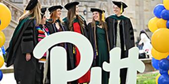 new graduates pose in caps and gown behind large Pitt script logo display and blue and gold balloon towers.