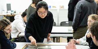 instructor points out elements of a piece of artwork lying on the table to students on either side.