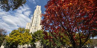 Cathedral of Learning stands behind leafy trees starting to change color in autumn with wispy clouds against blue sky in the background.